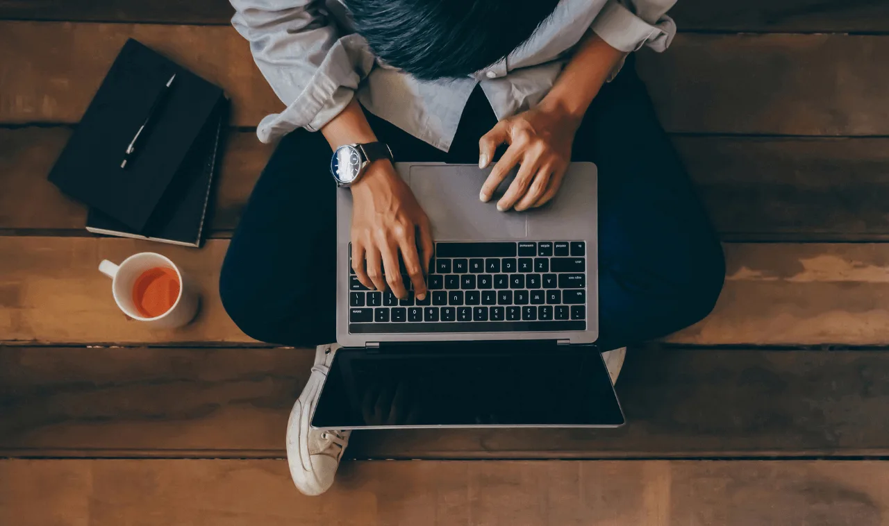 Man sitting on the floor on his computer Man sitting on a wooden floor on his computer with a cup of tea and a notebook sitting on the floor next to him.