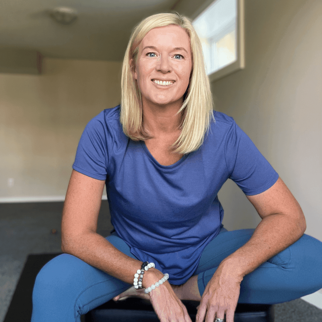 Image of Sinead Oconnor sitting on a chair in a room posing for a professional photo. She is a blonde, white woman wearing a blue t shirt and blue leggings.